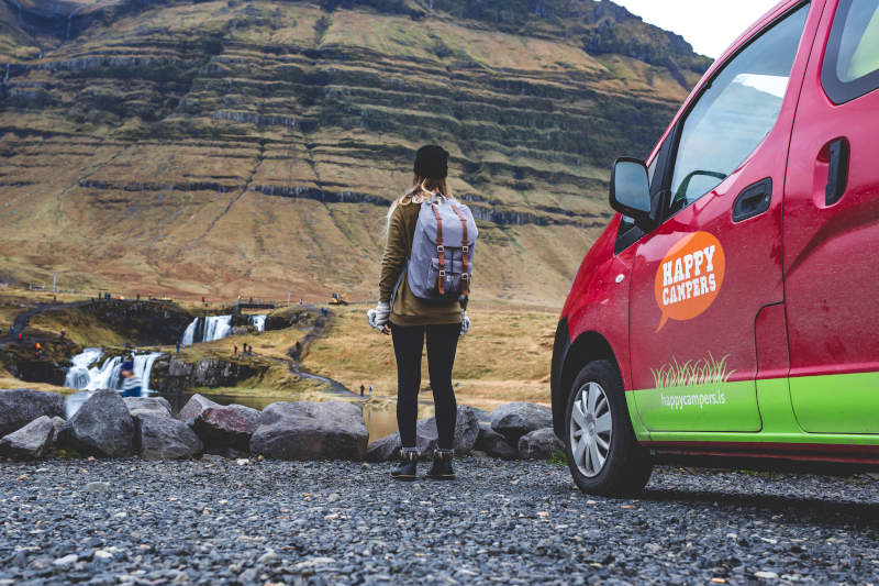 A lady with a backpack standing next to a red campervan at the foot of the mountain. 