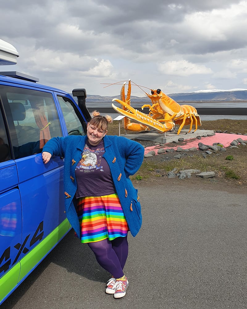 a woman with a blue sweater and colorful skirt standing next to a blue campervan in front of the giant plastic yellow lobster