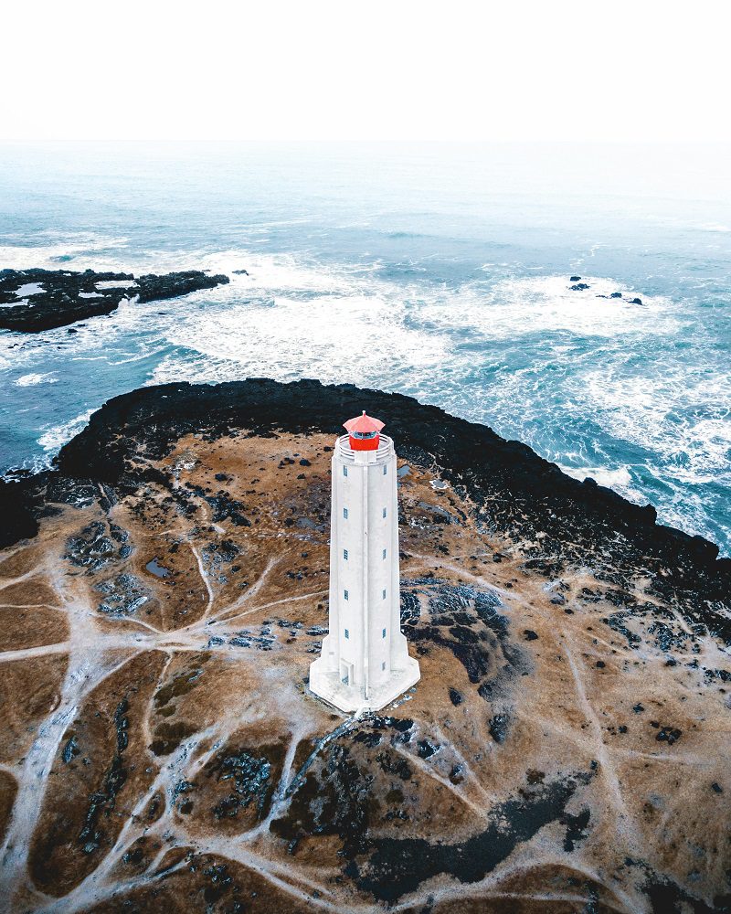 Arial shot of stark white Malariff lighthouse with bright red tower top, surrounded by dramatic Icelandic landscape of black lava cliffs and Atlantic Ocean