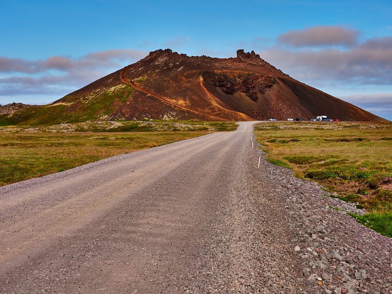 A photo taken from a gravel road leading towards redddish-brown cone shaped Saxholl crater in Snaefellsnes Nation Park