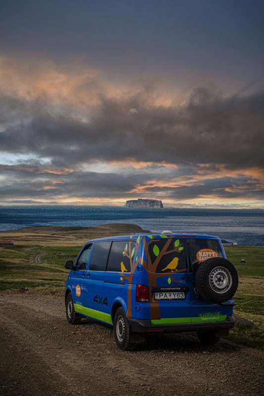 Happy Campers 4x4 campervan parked on a scenic gravel road in Iceland, overlooking the ocean and dramatic cliffs at sunset.
