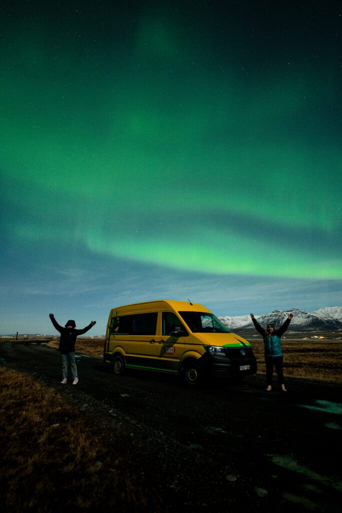 Two travelers standing with arms raised beside a yellow Happy Campers van under the Northern Lights in Iceland, with snow-capped mountains in the distance.

