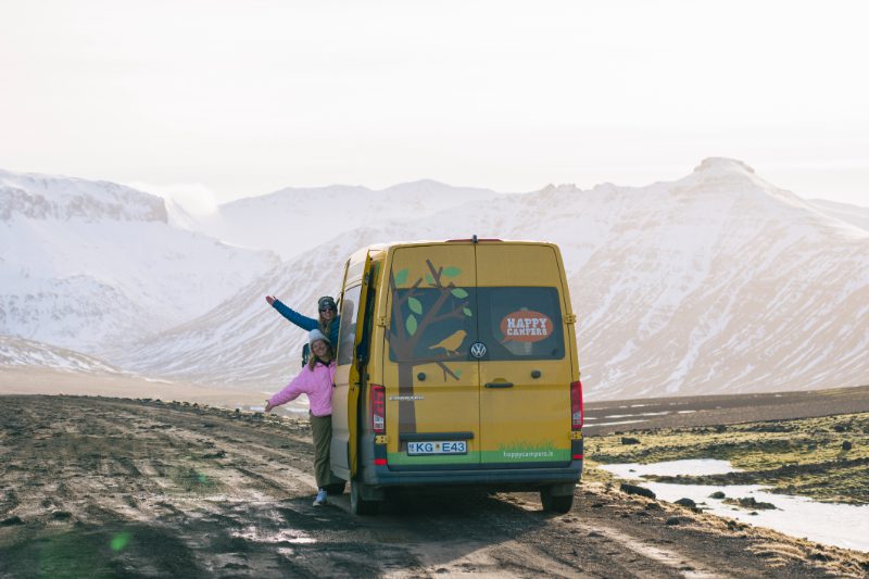 Two travelers posing at the back of a yellow Happy Campers van on a remote Icelandic road, surrounded by snow-capped mountains and early spring thaw