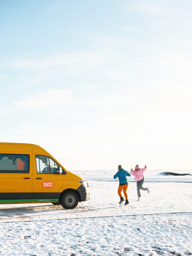 Two joyful travelers dancing in the snow beside a yellow Happy Campers van on a bright winter day in Iceland