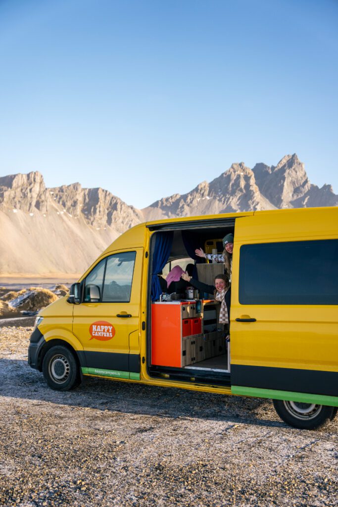 Two smiling travelers waving from inside a yellow Happy Campers van parked near Vestrahorn mountain in Iceland, with scenic rocky peaks in the background