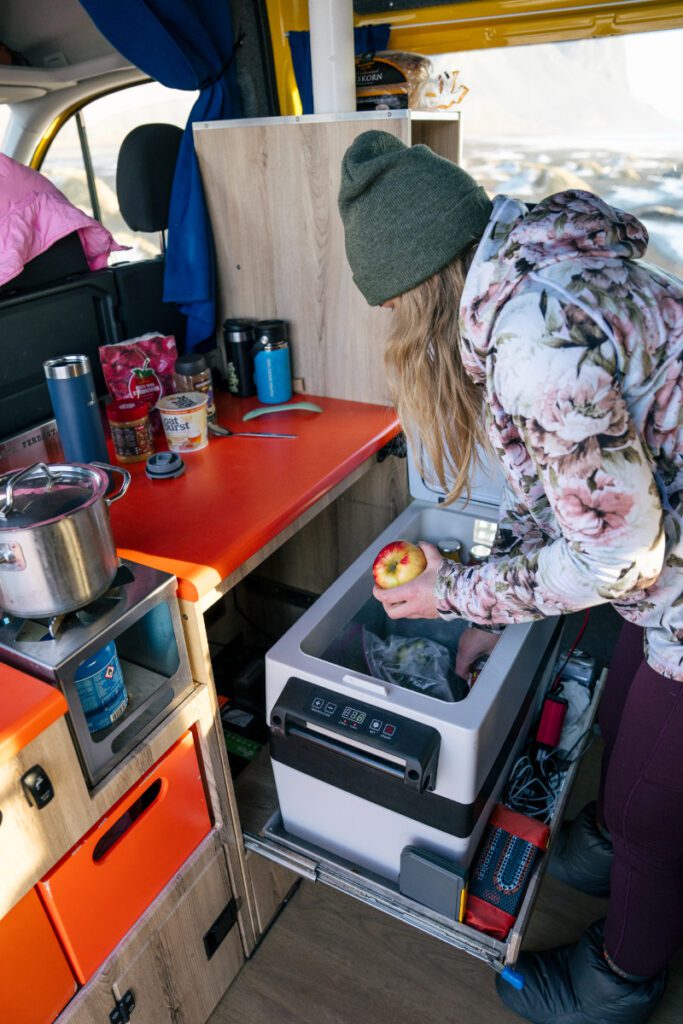 Traveler reaching into a campervan fridge to grab an apple inside a Happy Campers van, surrounded by cooking essentials and a gas stove setup