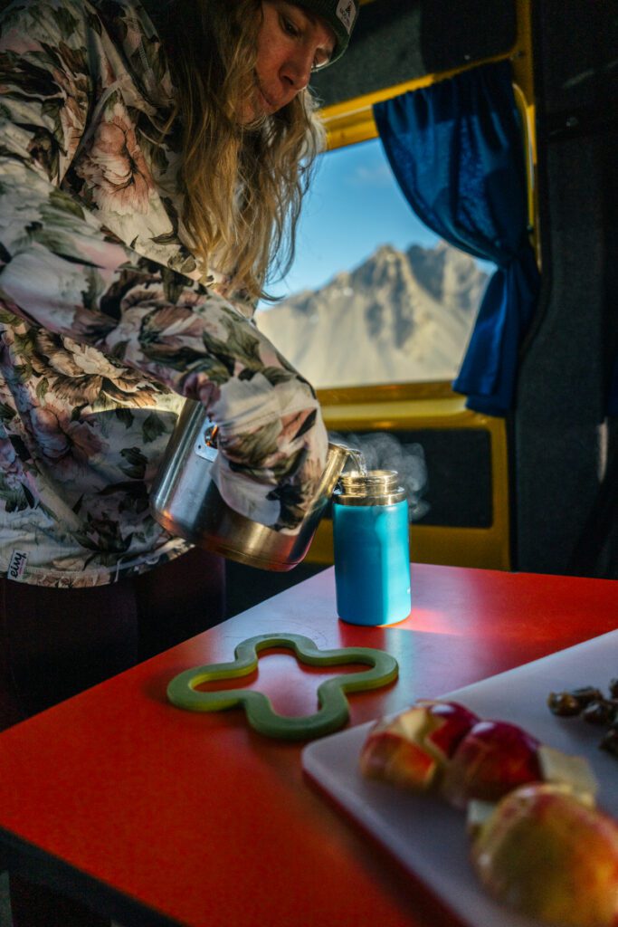 Traveler pouring hot water into a thermos inside a campervan with mountain views through the window, next to sliced apples and snacks on the table