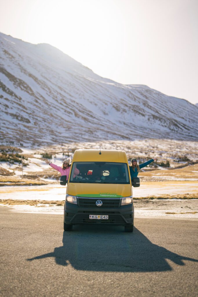 Two smiling travelers leaning out of a yellow Happy Campers van with arms outstretched, driving through a snowy Icelandic mountain valley in winter sun