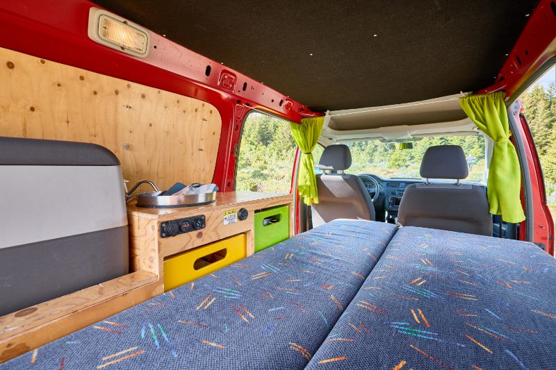 Happy 1 EX AUTO campervan rental interior in Iceland, featuring a cozy double bed, compact kitchenette with fridge and drawers, and a scenic view through the front cabin.