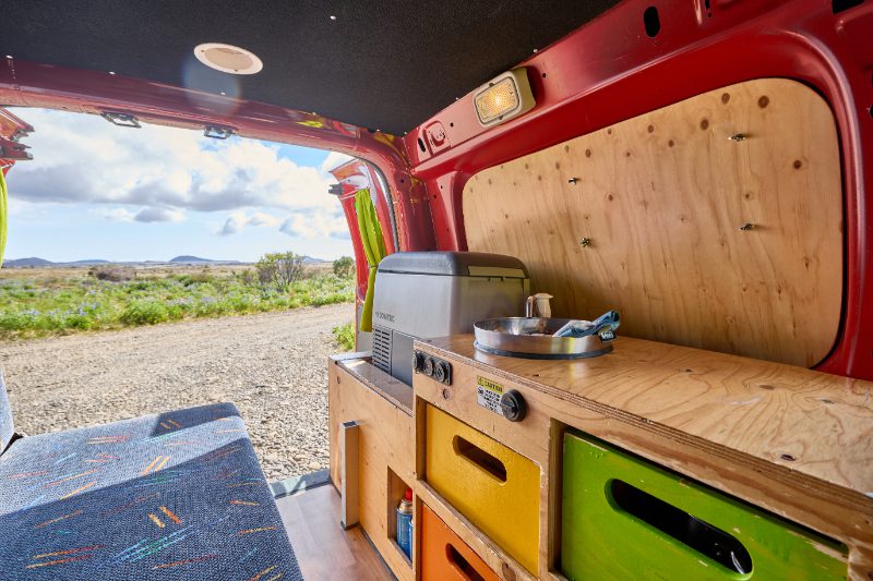 Compact kitchenette inside a Happy Campers van rental in Iceland, featuring colorful storage drawers, a portable stove, and mini fridge with a scenic outdoor view.