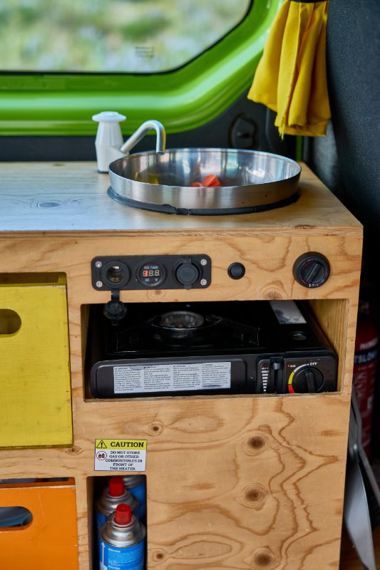 Compact kitchen setup in the Happy 2 EX campervan rental in Iceland, featuring a portable gas stove, sink, and colorful storage drawers—perfect for off-grid cooking on the road.