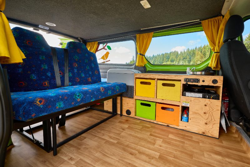 Interior of the Happy 2 EX campervan showing colorful storage drawers, a compact kitchenette with sink and stove, a fridge, and a rear bench seat with safety belts.