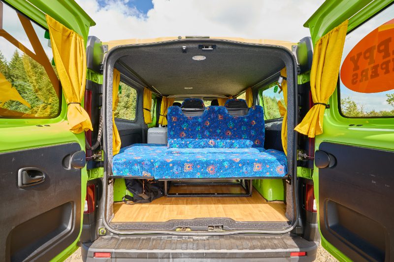 Rear view of the Happy 2 EX campervan showing the two-person bench seat, vibrant yellow curtains, and wood-style flooring.