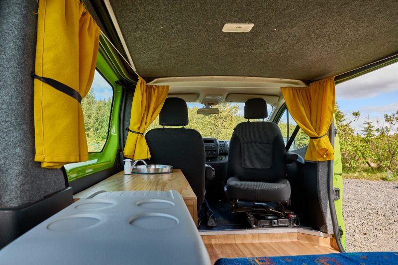 View from the back of the Happy 2 EX campervan showing the front seats, compact kitchenette with sink, and yellow curtains on large windows.