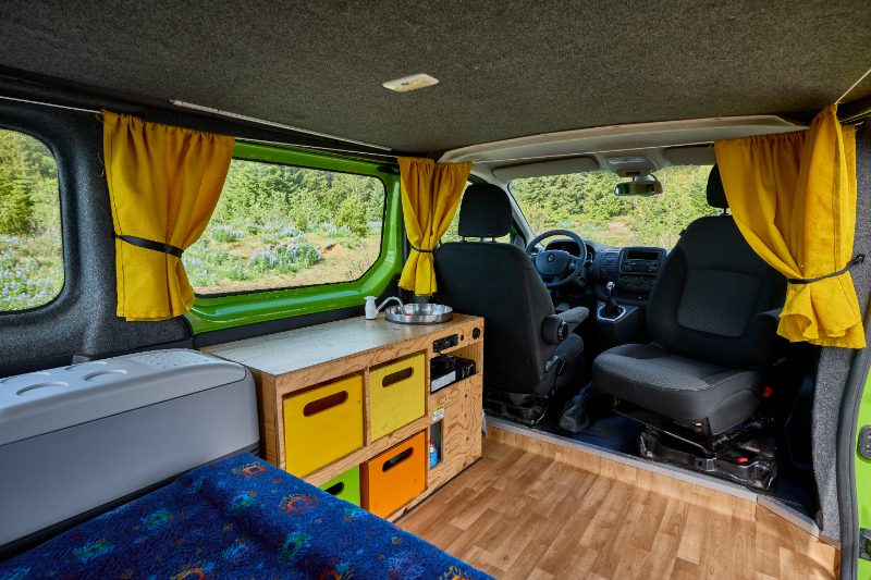 Interior of the Happy 2 EX campervan featuring the front seats, wooden kitchenette with colorful storage, and a compact fridge under large windows with yellow curtains.