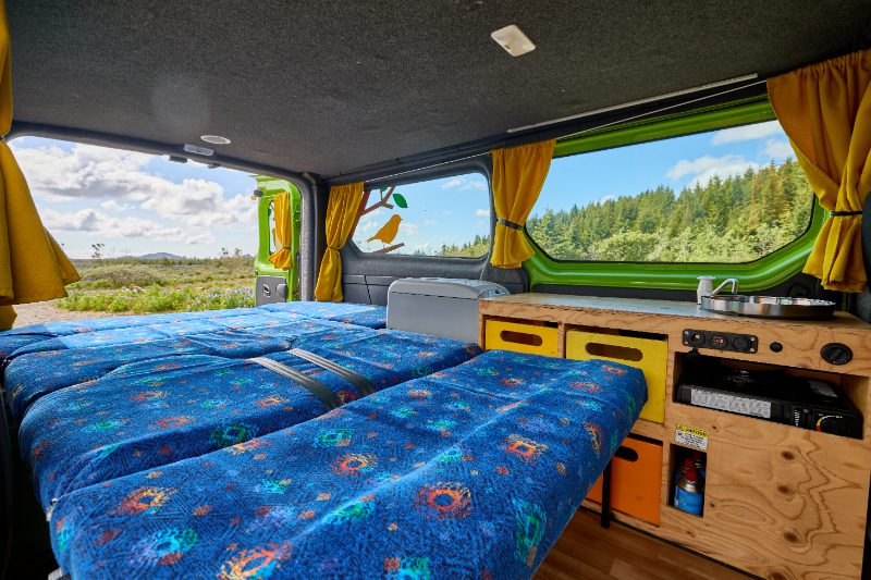 Interior of the Happy 2 EX campervan rental in Iceland, showcasing a fully extended bed, built-in kitchenette, and panoramic windows with forest views—ideal for two-person vanlife comfort.