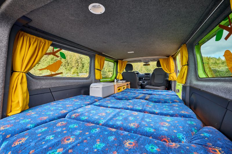 Wide-angle interior view of the Happy 2 EX campervan rental in Iceland, featuring a spacious double bed setup, yellow curtains, and a compact kitchenette—perfect for cozy van travel.