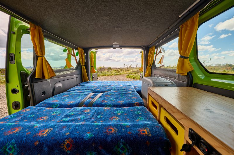 Rear interior view of the Happy 2 EX campervan rental in Iceland, with a double bed setup, yellow curtains, kitchenette, and open back doors revealing Iceland’s natural beauty.